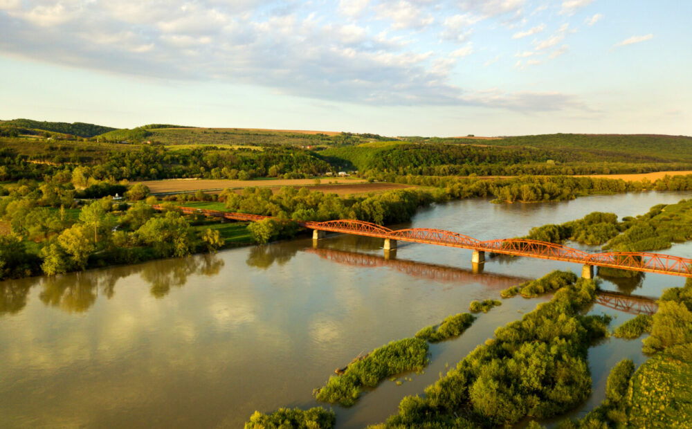Aerial view of a narrow road bridge stretching over muddy wide river in green rural area.